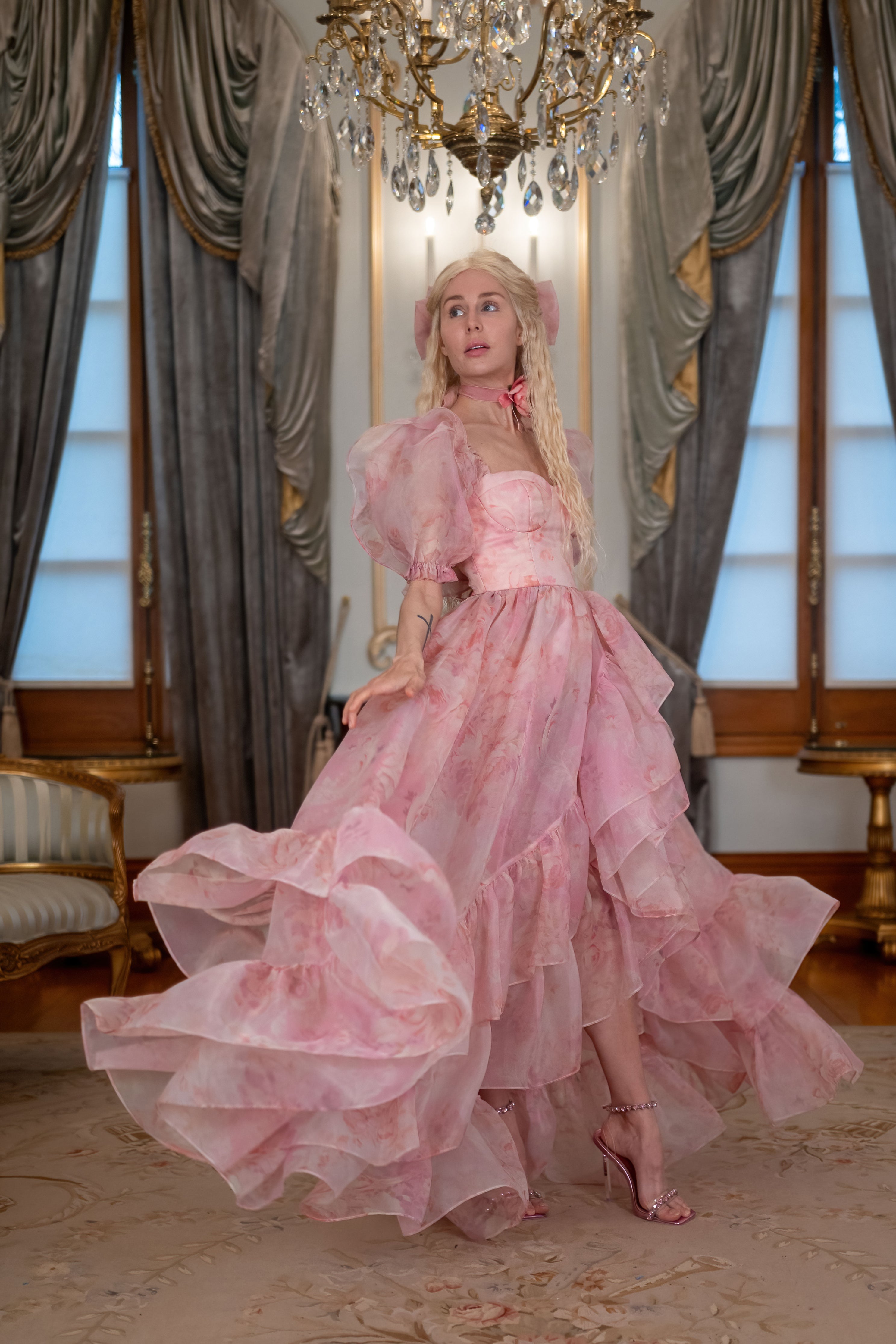 A woman wearing a pink, floral, maxi dress with a corseted back and a thigh-high split, standing in a luxurious room with a chandelier.