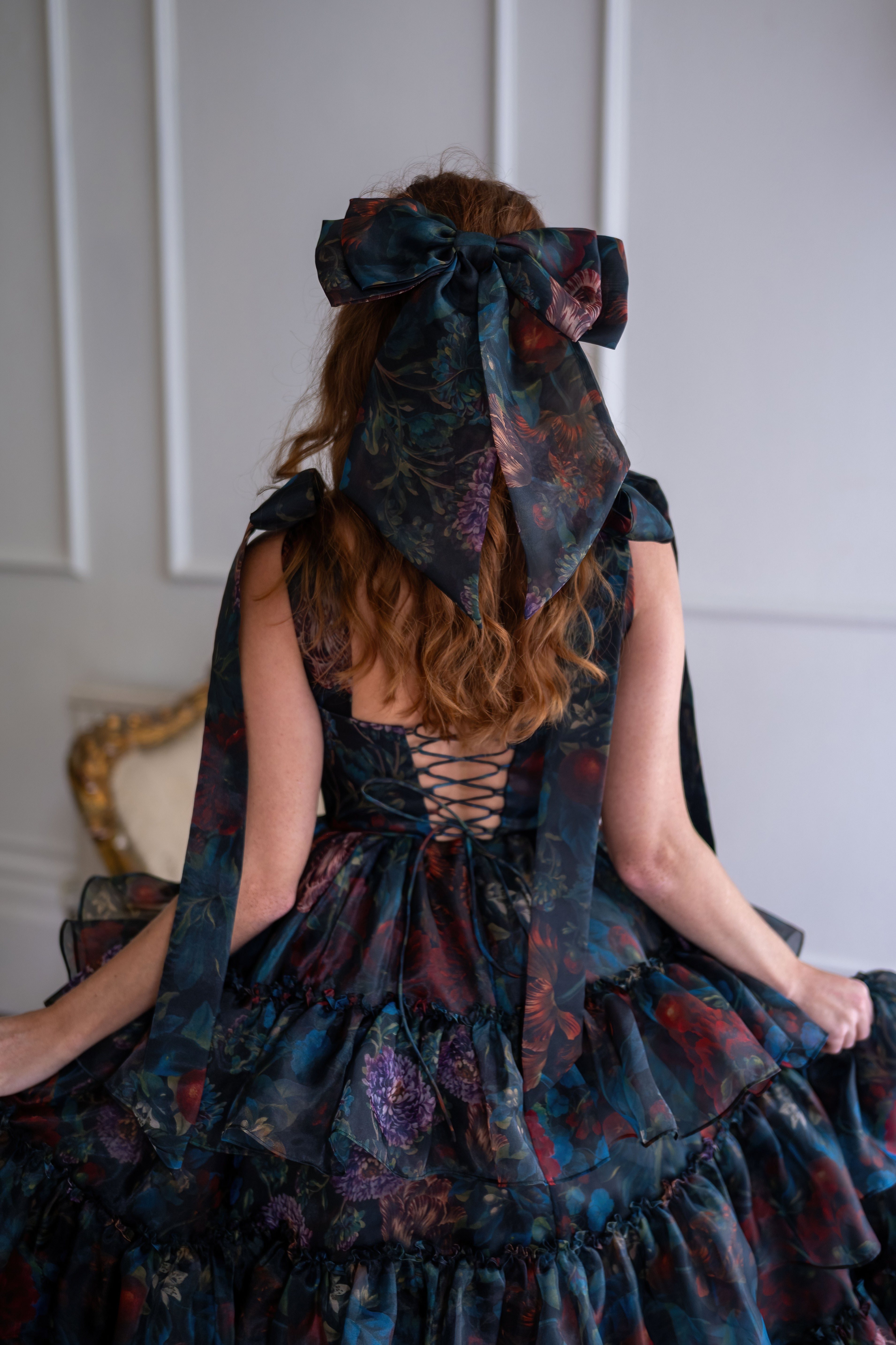 Person wearing a dark, patterned dress with a large bow on their head against a neutral background